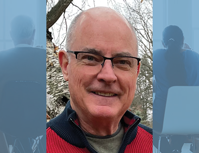 Smiling man with glasses outdoors, wearing a red jacket, bare trees in the background.