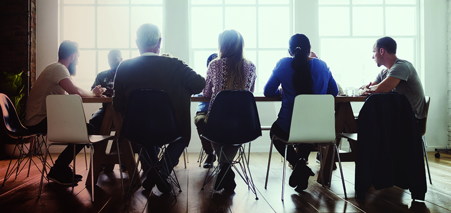Back view of a diverse group seated around a wooden conference table in a bright, window-filled office space.