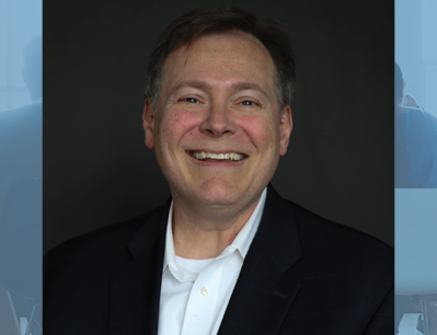 Portrait of a smiling man in a dark suit jacket and white shirt against a dark background, head and shoulders visible.