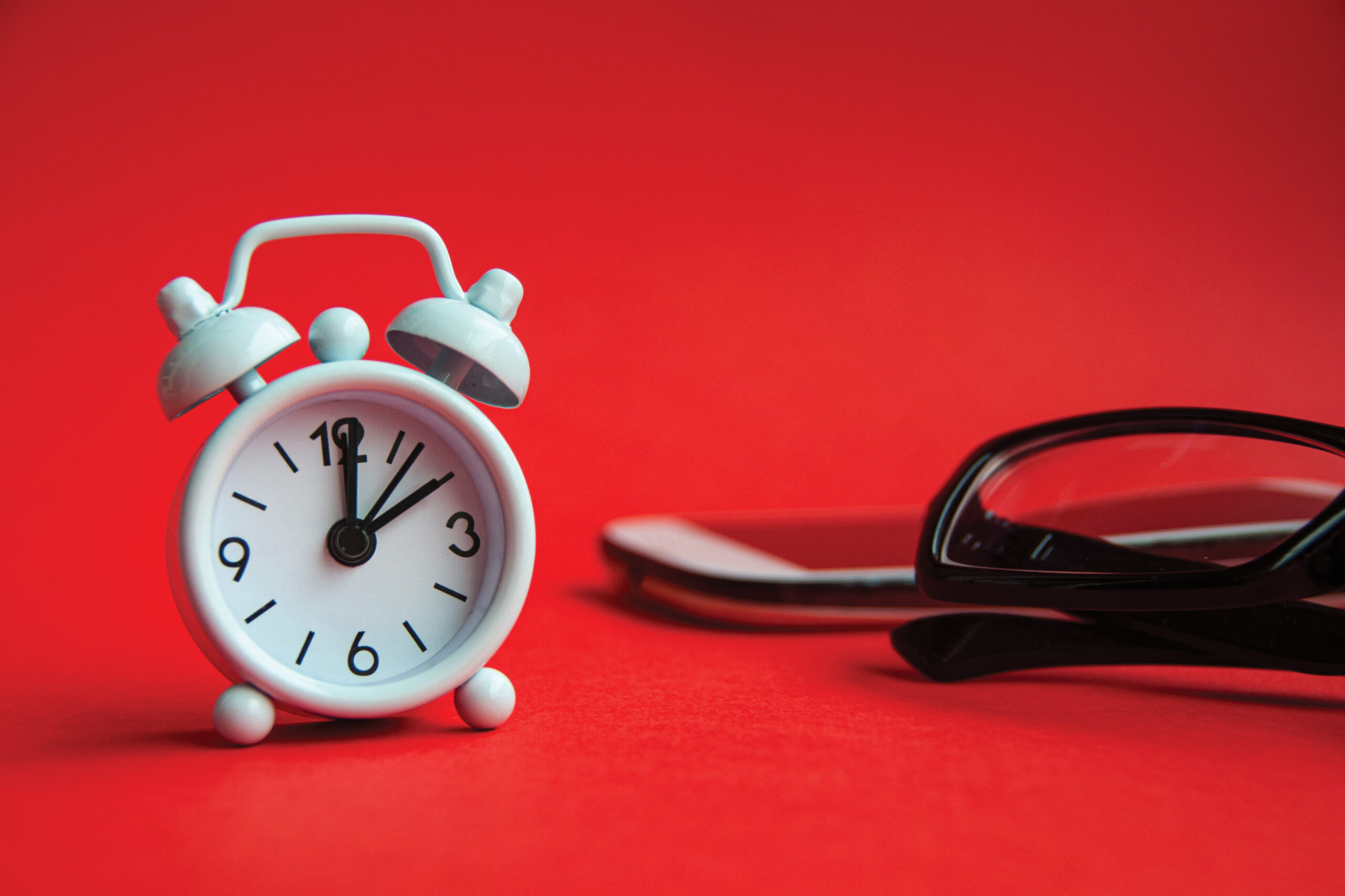 White alarm clock with bells on a red surface beside black-framed glasses.