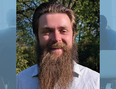Bearded man with light brown hair smiling outdoors, trees and sunlight in the background