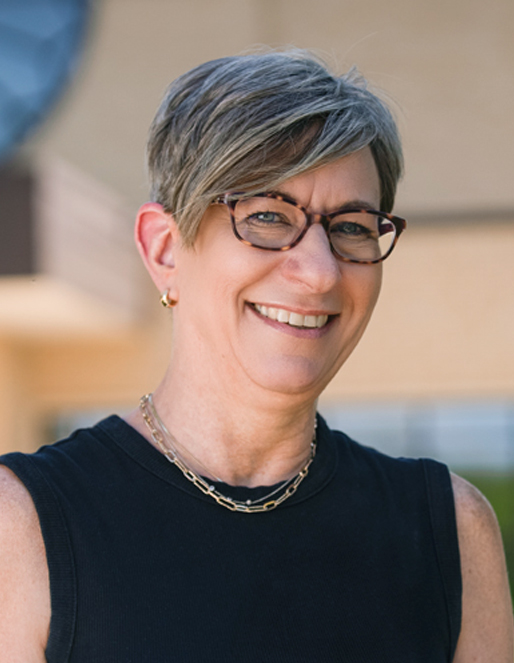 Smiling woman with short gray hair and glasses in a sleeveless black top outdoors, facing the camera.