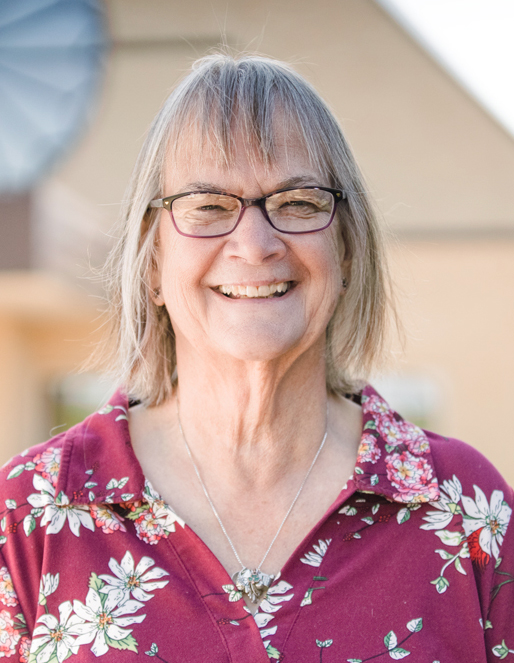 Smiling elderly woman wearing glasses and a floral burgundy shirt, outdoors, with a blurred building in the background.