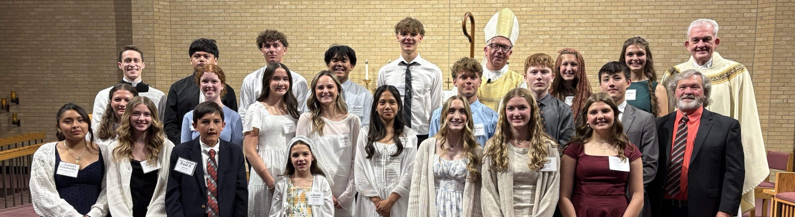 Group portrait of teens and adults in formal attire on church steps, with a bishop in mitre in the background.