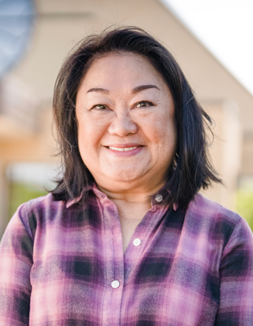 Smiling woman with shoulder-length dark hair wearing a purple plaid shirt, outdoors with a blurred background.