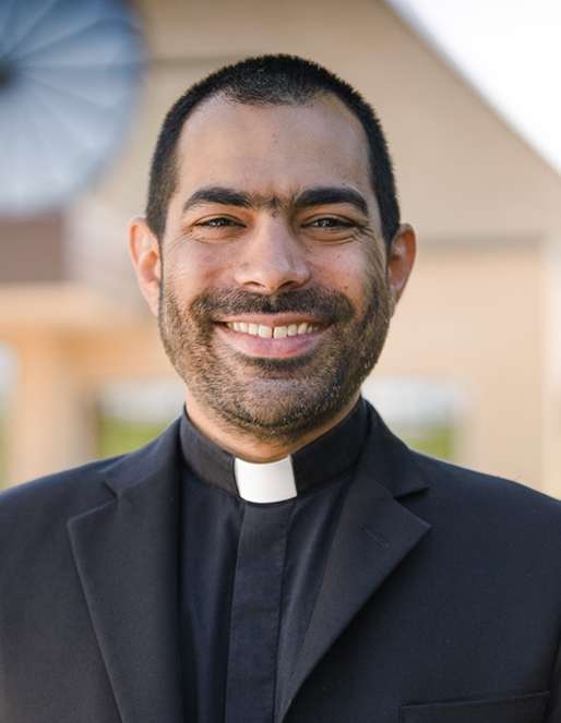 Smiling man in a black suit with a white clerical collar, standing outdoors.