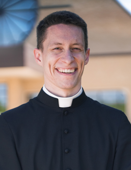 Smiling priest wearing a black clerical shirt with a white collar outdoors at a sunny location.