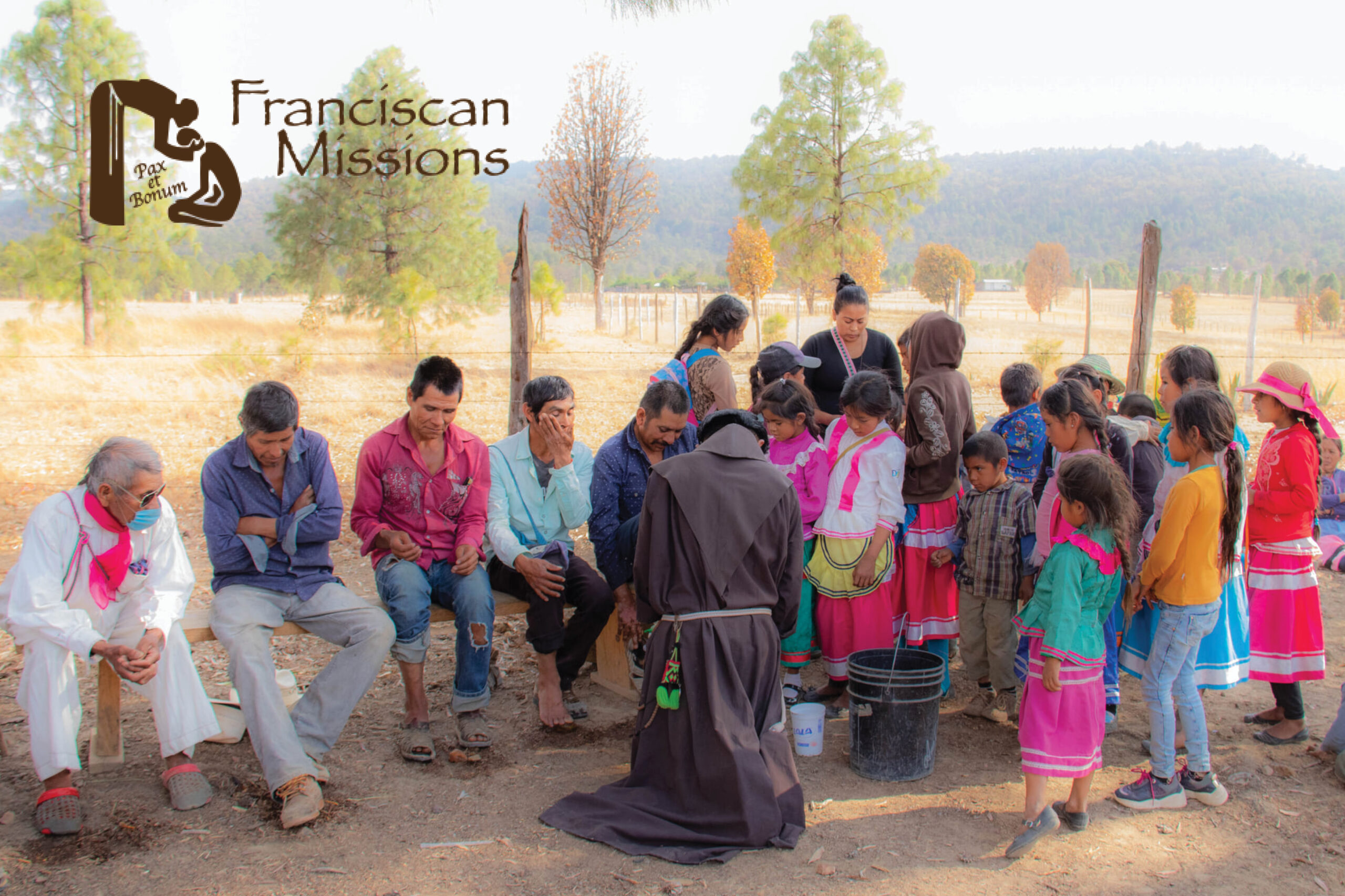 Community gathering outdoors in a rural setting, children in colorful dress around adults; a woman in dark robes leads a ritual near a bucket, with a Franciscan Missions logo on the left.
