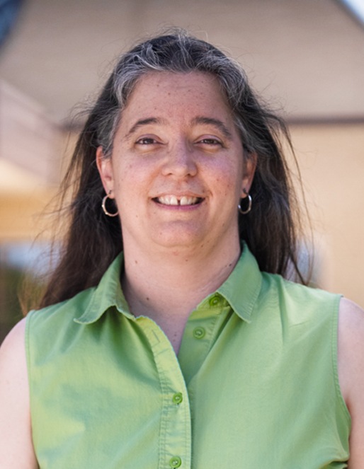 Smiling woman with shoulder-length dark hair, wearing a lime green sleeveless shirt and hoop earrings, outdoors on a sunny day.