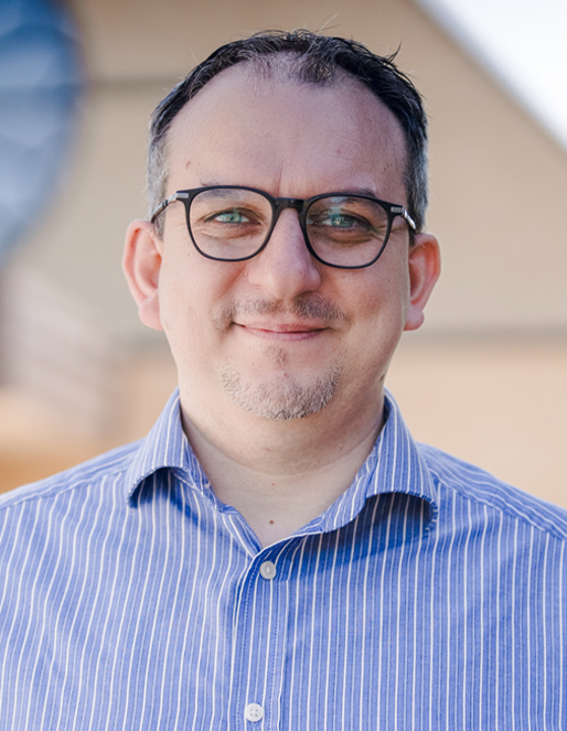 Portrait of a man with short dark hair and glasses wearing a blue striped button-down shirt, smiling outdoors.