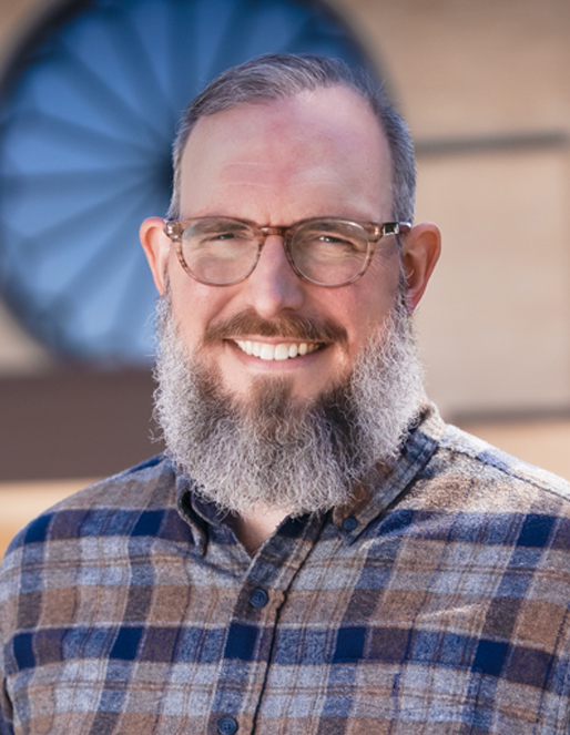 Portrait of a smiling man with a gray beard and round glasses wearing a blue plaid shirt outdoors.