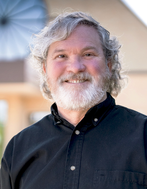 Smiling older man with gray hair and beard in a black button-down shirt, outdoors.