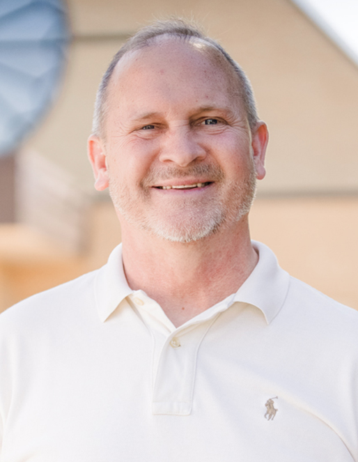 Smiling man in a white polo shirt outdoors, neutral background behind him.