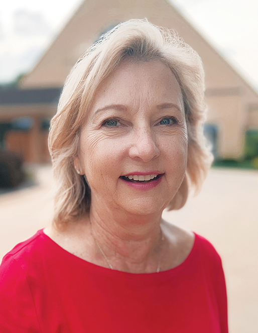 Smiling woman with short blonde hair wearing a red top, standing outdoors with a building in the background.