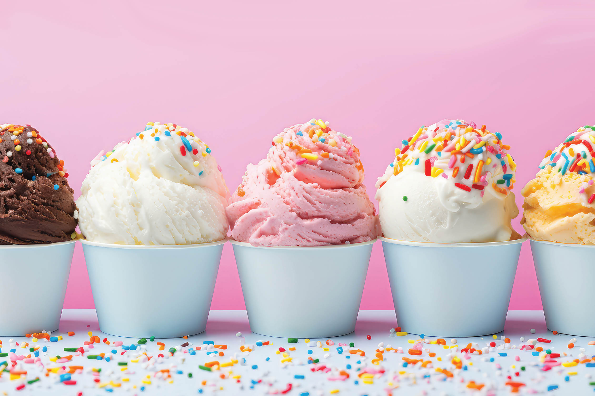 Row of colorful ice cream scoops with rainbow sprinkles in light blue cups against a pink background, festive and bright.