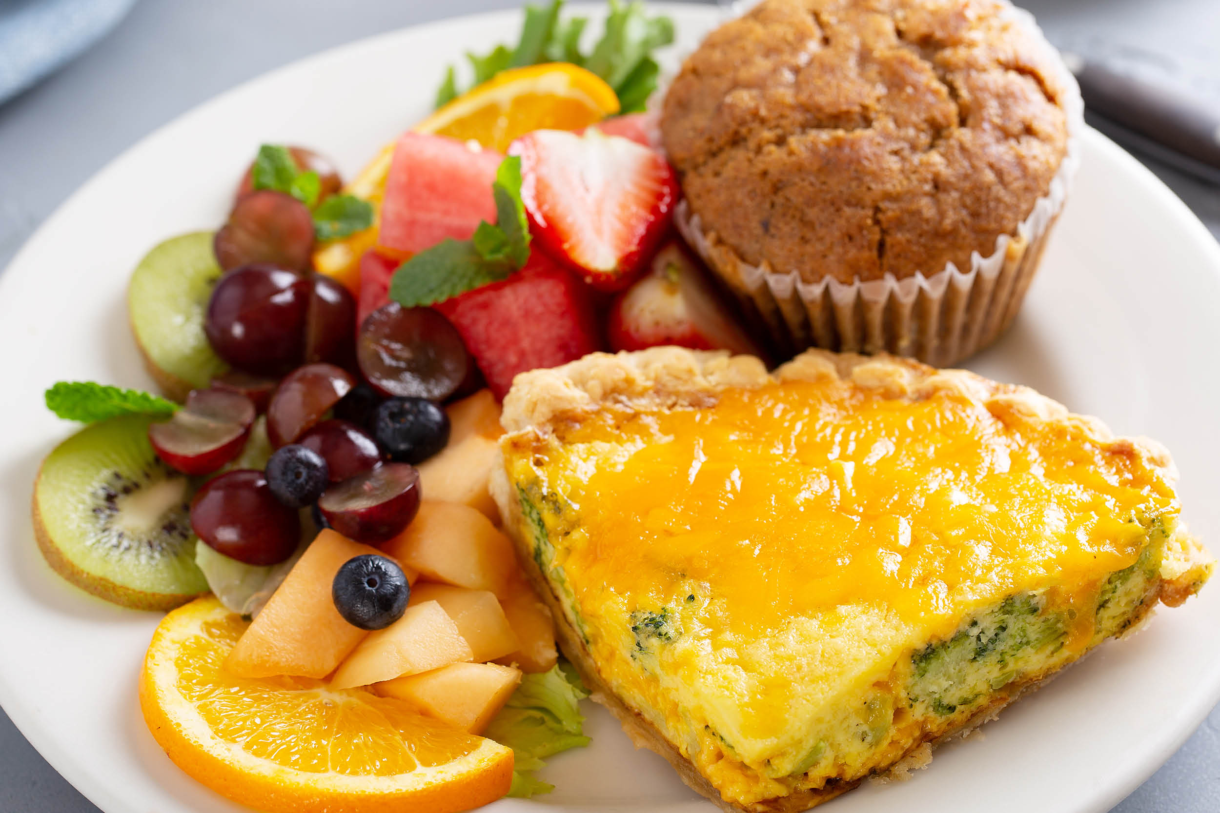 Fruit plate with orange wedges, kiwi, grapes, blueberries, strawberry, mint, plus a savory muffin and a cheese-spinach quiche slice on a white plate