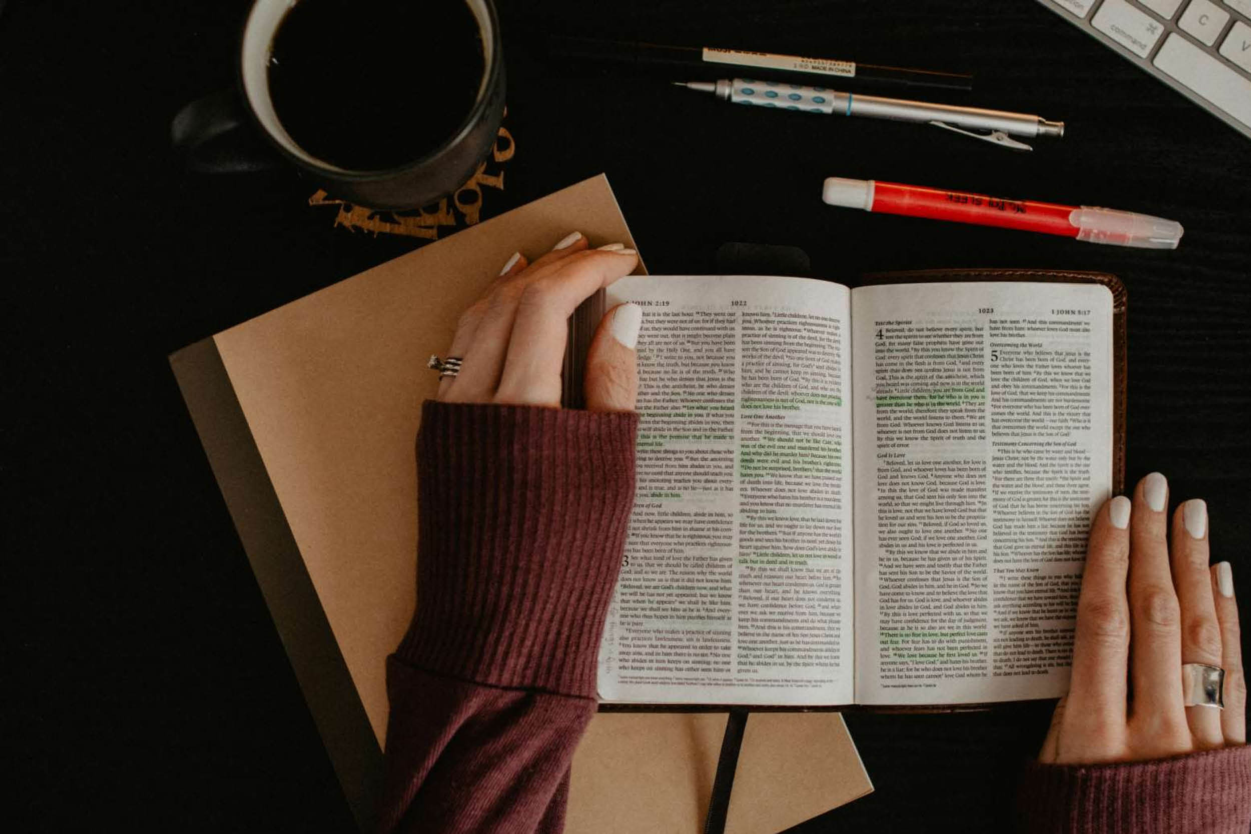 Open Bible on a dark desk with a person turning pages, accompanied by a mug, notebook, and pens.