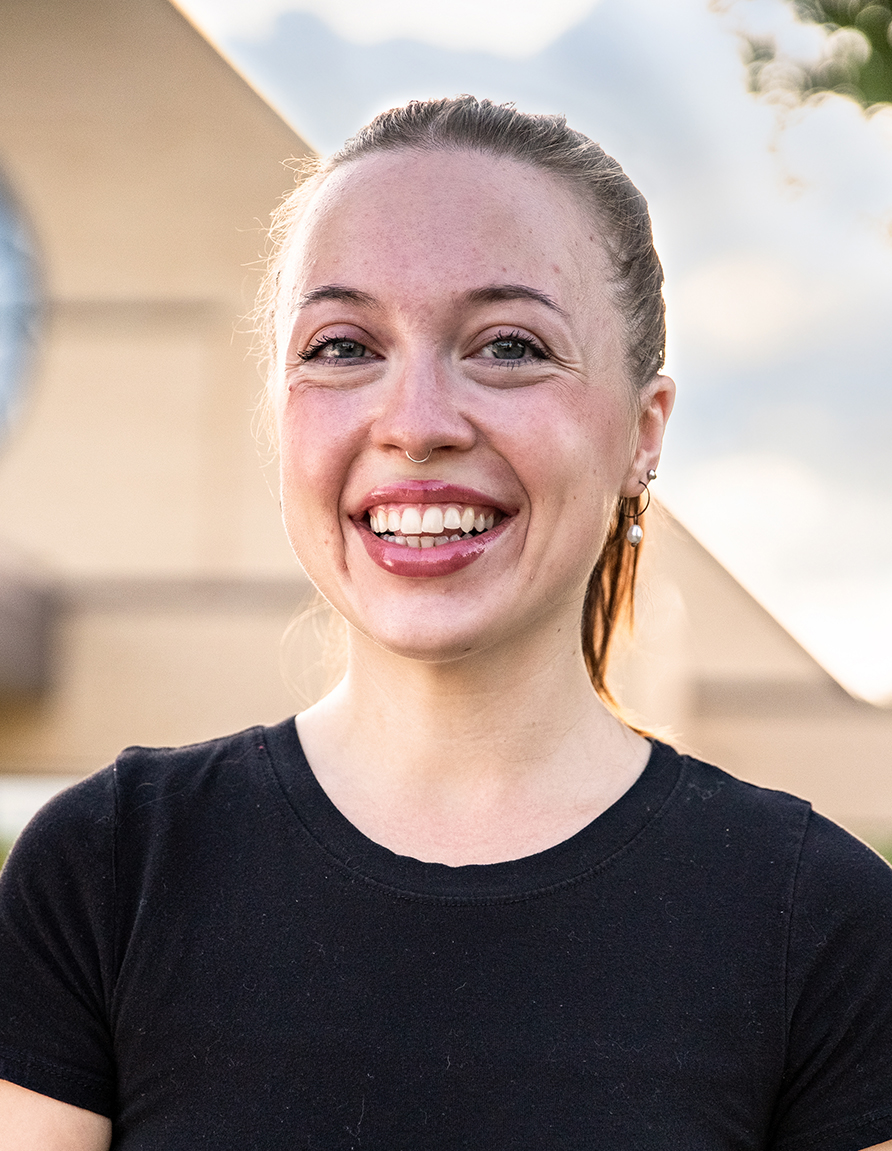 Smiling woman with a nose ring in a black shirt, outdoors with a blurred building in the background.