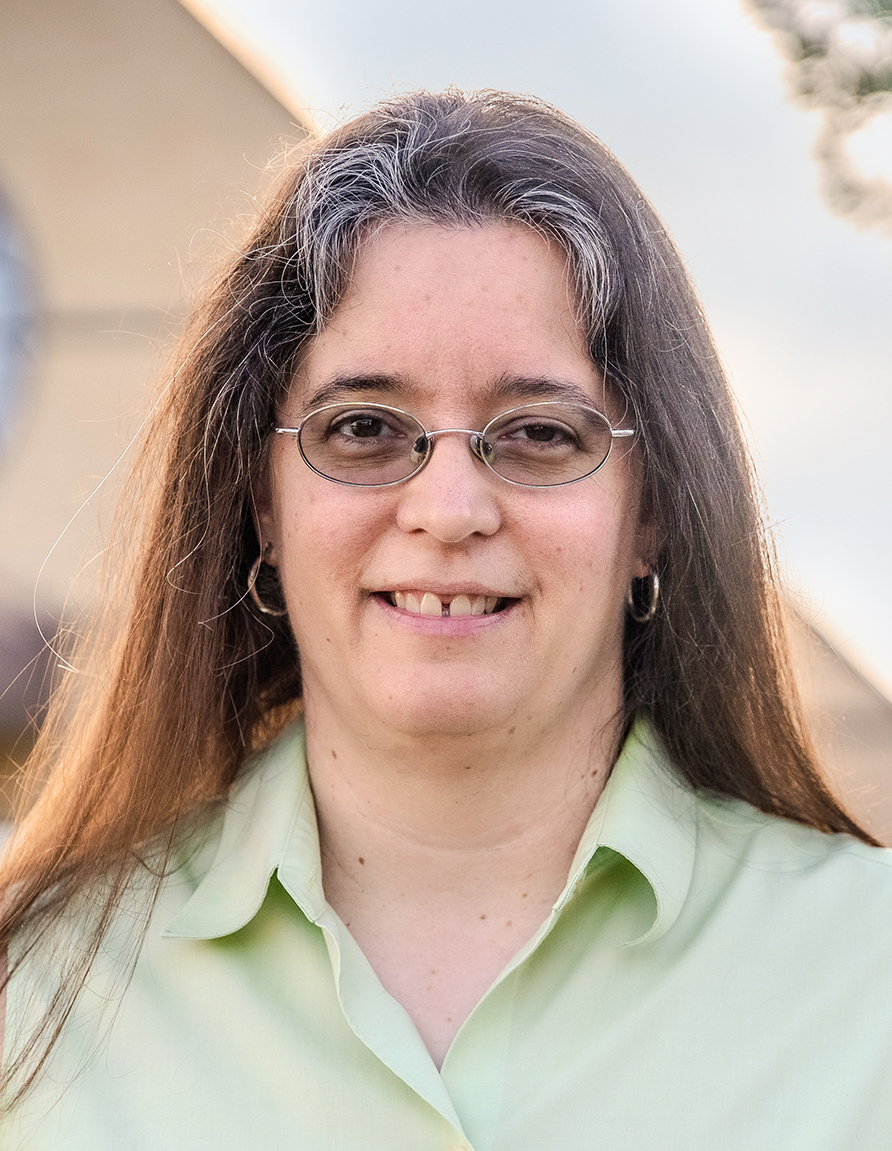 Woman with long brown hair and glasses smiling outdoors, wearing a light green collared shirt.