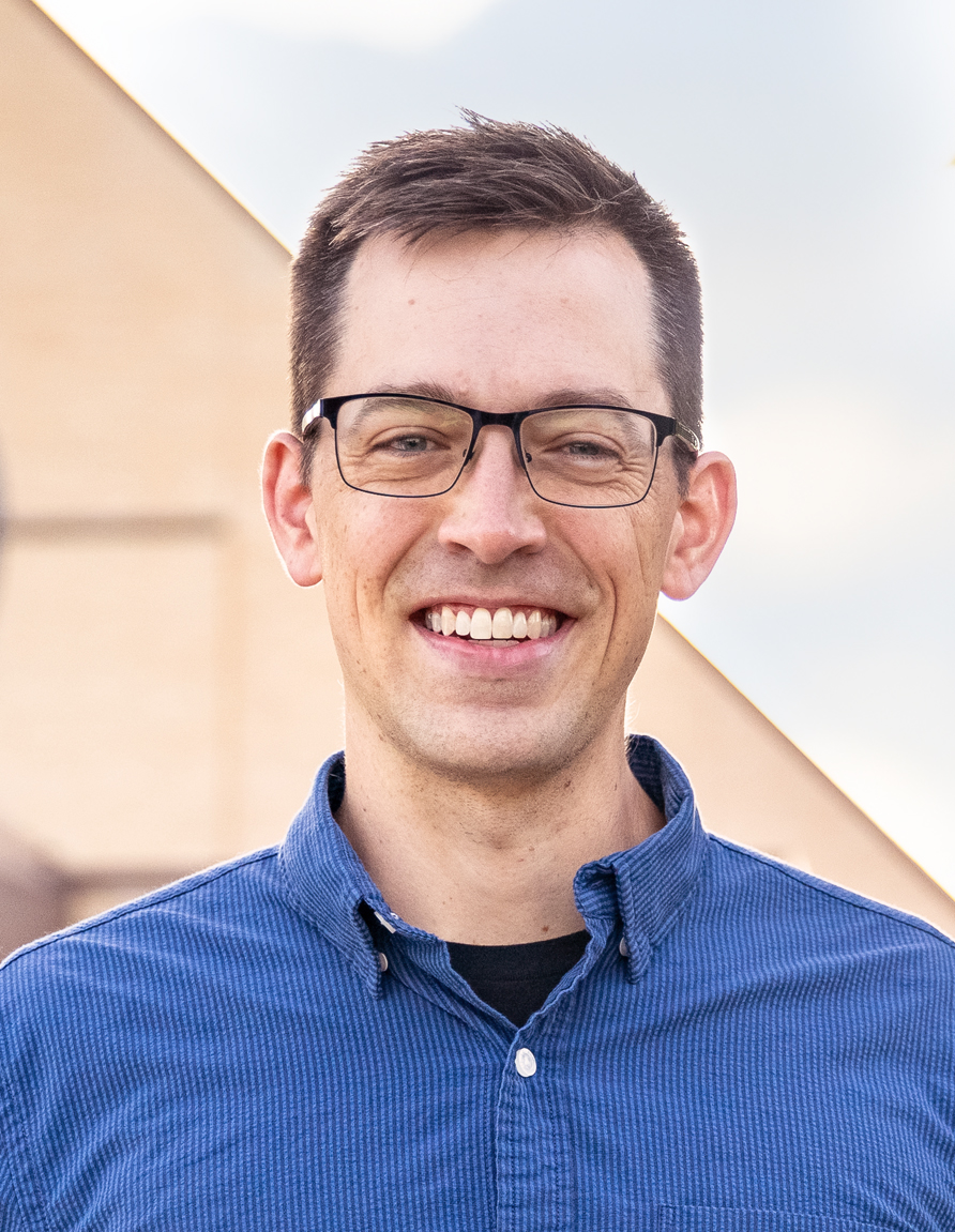 Smiling man with short dark hair and glasses wearing a blue button-down shirt outdoors.